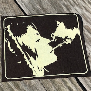 May include: A rectangular patch featuring a profile of a person exhaling smoke. The image is in a black and cream color scheme, with a white border. The patch is likely made of fabric and is suitable for sewing or ironing onto clothing or accessories.