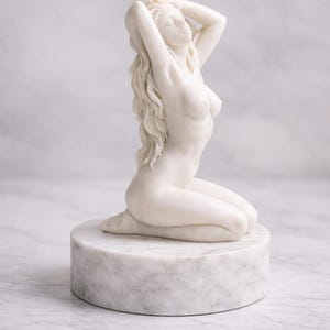 Puede incluir: Una vela de cera blanca con forma de figura arrodillada de cabello largo y fluido, colocada sobre una base circular de m&aacute;rmol blanco. La vela tiene una mecha encendida en la parte superior de la cabeza, proyectando una llama sobre la superficie.