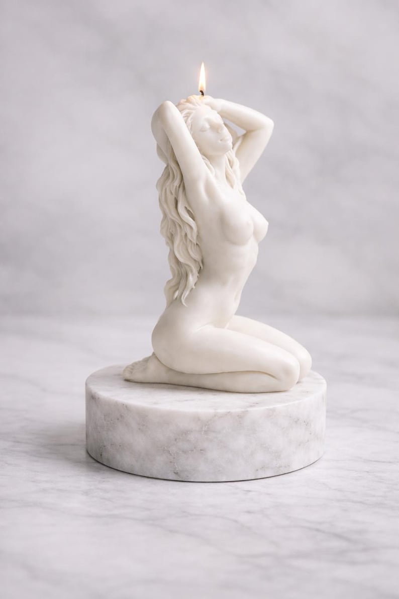 Puede incluir: Una vela de cera blanca con forma de figura arrodillada de cabello largo y fluido, colocada sobre una base circular de m&aacute;rmol blanco. La vela tiene una mecha encendida en la parte superior de la cabeza, proyectando una llama sobre la superficie.