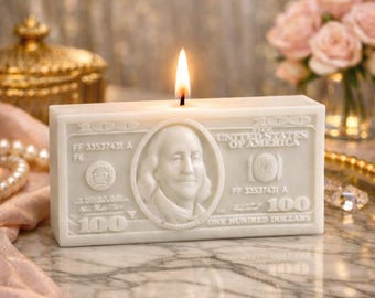 Puede incluir: Una vela blanca con forma de billete de 100 dólares, con una llama encendida. La vela presenta un retrato de Benjamin Franklin y el texto "UNITED STATES OF AMERICA".