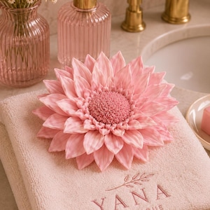 May include: A light pink, folded cotton towel featuring the embroidered text YANA BOTANIQUE with a small leaf icon. Resting on top is a large, detailed pink flower-shaped soap or decorative object with textured petals and a dense centre.