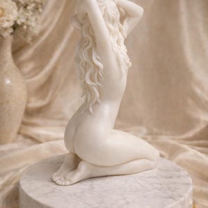 Puede incluir: Una vela de cera blanca esculpida con la forma de una figura arrodillada con cabello largo y fluido. La figura se muestra con los brazos levantados, las manos tocando la nuca, y se asienta sobre una base circular de m&aacute;rmol blanco con borde dorado.