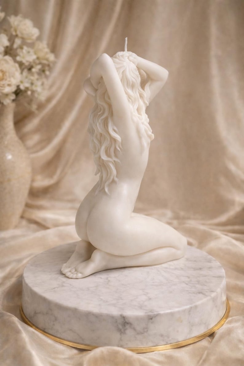 Puede incluir: Una vela de cera blanca esculpida con la forma de una figura arrodillada con cabello largo y fluido. La figura se muestra con los brazos levantados, las manos tocando la nuca, y se asienta sobre una base circular de m&aacute;rmol blanco con borde dorado.