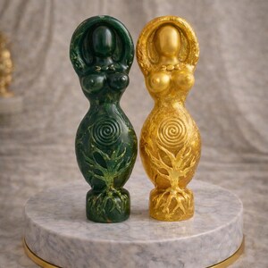 May include: Two goddess statues, one green and one gold, stand on a marble pedestal with a gold trim. The statues have a tree of life design on the lower half and a spiral design on the upper half. The statues are approximately 6 inches tall.