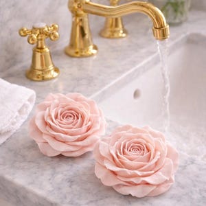 May include: Two light pink rose-shaped soaps rest on a marble countertop next to a gold faucet with running water. A white towel and a vase of pink roses are also visible.