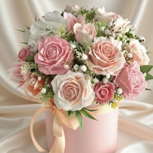 May include: A round, light pink gift box overflowing with a bouquet of roses in various shades of pink and white. The arrangement is accented with small white flowers and greenery, tied with a peach ribbon.