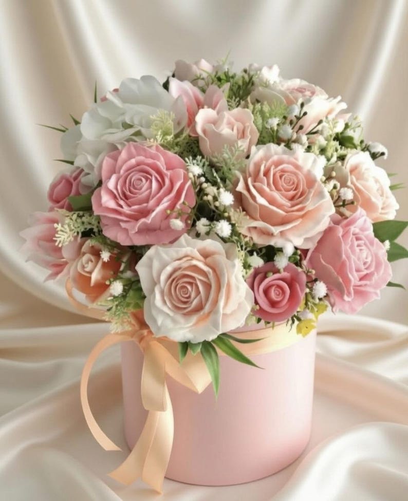 May include: A round, light pink gift box overflowing with a bouquet of roses in various shades of pink and white. The arrangement is accented with small white flowers and greenery, tied with a peach ribbon.