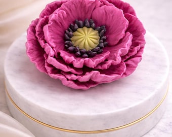 May include: A vibrant magenta flower with layered petals, featuring a yellow and dark center. The flower rests on a white, round marble box with a gold trim. The background includes a soft, white fabric.
