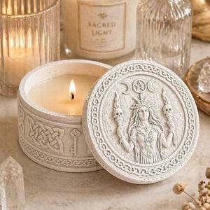 Può includere: Una candela accesa in un contenitore rotondo bianco con un coperchio dettagliato con un disegno scolpito di una donna, lune e teschi. Il contenitore è circondato da altre candele e oggetti decorativi.