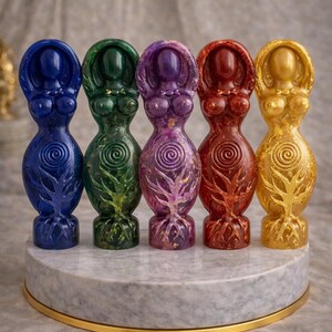 May include: Five colorful resin goddess statues in blue, green, purple, red, and gold. Each figure has a stylized form with a tree-like design at the base and a spiral on the torso. They are displayed on a round, marble-like platform.