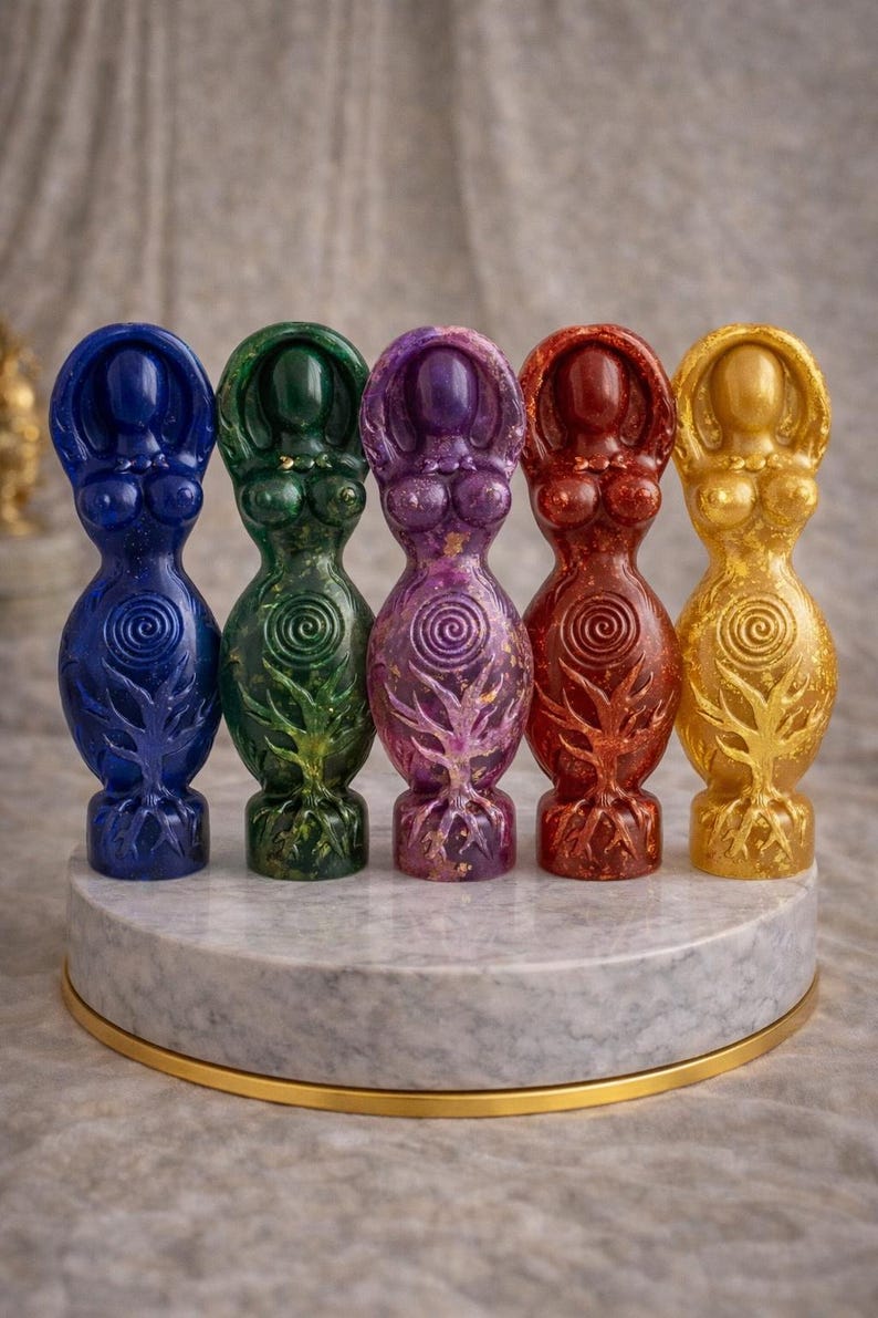 May include: Five colorful resin goddess statues in blue, green, purple, red, and gold. Each figure has a stylized form with a tree-like design at the base and a spiral on the torso. They are displayed on a round, marble-like platform.