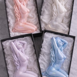 Puede incluir: Cuatro velas decorativas con forma de figuras arrodilladas de cabello largo y fluido, presentadas cada una en una caja rectangular negra forrada con papel de seda blanco. Las velas tienen acabados en colores rosa pastel, blanco, lavanda y azul claro.