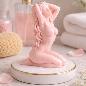 Puede incluir: Una vela de cera rosa p&aacute;lido con forma de figura arrodillada de largo cabello ondulado y brazos elevados sobre la cabeza. La vela se asienta sobre una base circular de cer&aacute;mica blanca con un fino borde de color dorado en la parte superior.
