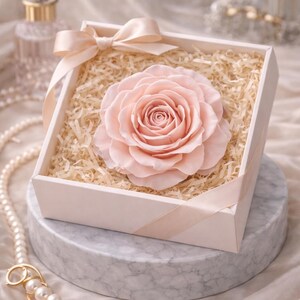 May include: A large, pale pink rose in a white gift box with a matching ribbon. The rose is surrounded by shredded paper. The box sits on a marble-like surface, with a pearl necklace and perfume bottle in the background.