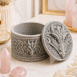 May include: A grey trinket box with a dragon design on the lid and sides. The lid is partially open. The box features Celtic knotwork and dragon head details. The text "PCL" is visible. The box is a decorative item.