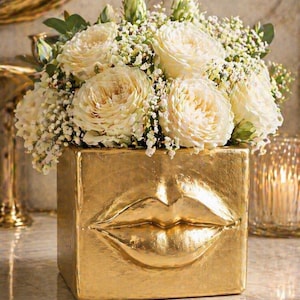 Puede incluir: Un jarrón cuadrado dorado con diseño de labios, lleno de rosas blancas y pequeñas flores blancas. El jarrón tiene una superficie texturizada y está colocado sobre una superficie similar al mármol.