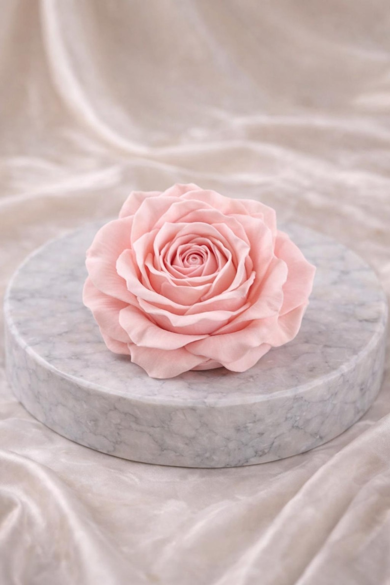 May include: A delicate, light pink rose in full bloom, centered on a round, marble-patterned platform. The rose's petals are soft and layered, with a tightly wound center. The platform sits on a draped, off-white fabric, creating a soft, elegant aesthetic.