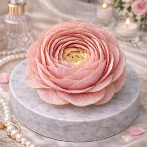 May include: A large, pink flower with gold trim sits atop a marble pedestal. The flower's petals are layered, creating a full, rounded shape. A pearl necklace and a perfume bottle are in the background, suggesting a theme of elegance and beauty.