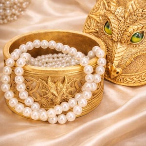 May include: A gold jewellery box with a dragon design, containing a pearl necklace. The box is open, showing more pearls within. A matching lid with a dragon face is beside it. The background is satin fabric.