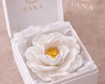 May include: A white peony flower with a yellow center rests inside a white box lined with tissue paper. The box is open, revealing the flower's delicate petals. The box has the gold text "YANA" and a "Y" logo.
