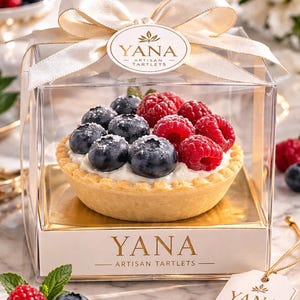 Puede incluir: Una sola tarta artesanal en una caja transparente, cubierta con arándanos frescos y frambuesas. La tarta está en una caja dorada con el nombre de la marca "YANA" impreso. También se incluye una cinta crema y una etiqueta con el nombre de la marca.