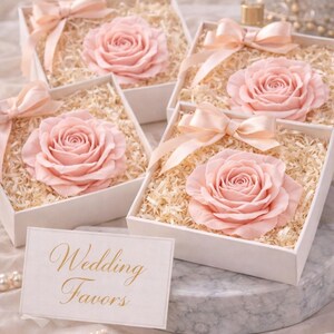 May include: Several white boxes filled with shredded paper and a large pink rose. Each box is tied with a light pink ribbon. A card with the words "Wedding Favors" is in the foreground. Pearls and a marble surface are also visible.