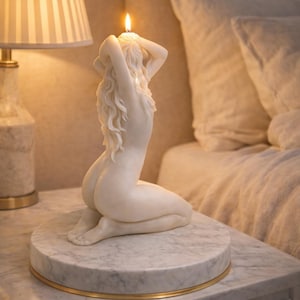 Puede incluir: Una vela de cera blanca con forma de figura arrodillada de largo cabello fluido, colocada sobre una base circular de m&aacute;rmol con borde dorado. La vela est&aacute; encendida, con una peque&ntilde;a llama que emerge de la parte superior de la cabeza.