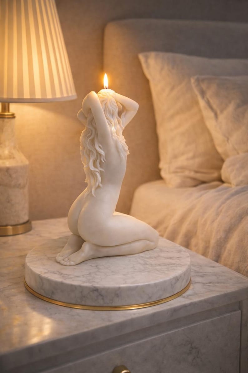 Puede incluir: Una vela de cera blanca con forma de figura arrodillada de largo cabello fluido, colocada sobre una base circular de m&aacute;rmol con borde dorado. La vela est&aacute; encendida, con una peque&ntilde;a llama que emerge de la parte superior de la cabeza.