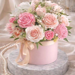May include: A round, light pink flower box overflowing with a bouquet of pink and white roses, accented with greenery. The box is tied with a peach ribbon and sits on a marble-patterned stand. A strand of pearls is draped nearby.