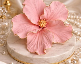 May include: A large, light pink artificial flower with ruffled petals and delicate gold-painted edges. The center of the flower features a textured, pale yellow stamen cluster. The flower is resting on a circular white marble base with a gold rim.