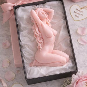 Puede incluir: Una vela de color rosa p&aacute;lido con forma de figura arrodillada de cabello largo y ondulado, presentada en una caja de regalo negra forrada con tela blanca. La caja tiene un lazo rosa y una etiqueta en forma de coraz&oacute;n que dice Happy Mother's Day.