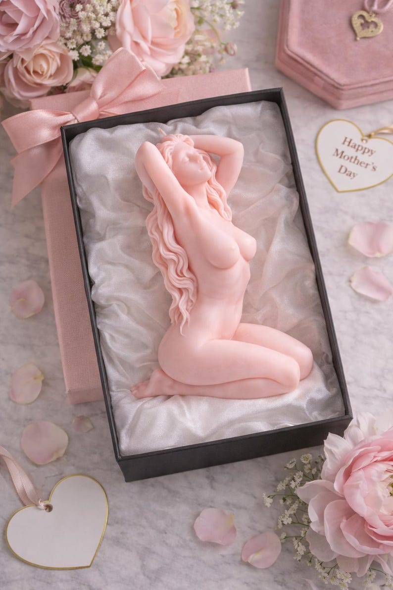 Puede incluir: Una vela de color rosa p&aacute;lido con forma de figura arrodillada de cabello largo y ondulado, presentada en una caja de regalo negra forrada con tela blanca. La caja tiene un lazo rosa y una etiqueta en forma de coraz&oacute;n que dice Happy Mother's Day.