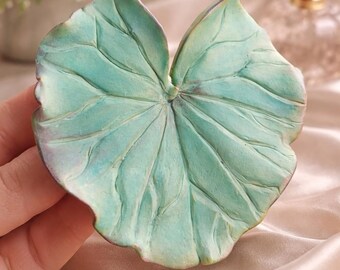 May include: A teal-coloured, leaf-shaped ceramic dish with detailed veining. The dish has a textured surface and a slightly aged appearance. The decorative leaf dish is likely for jewellery or small items. The dish is measured in centimetres.