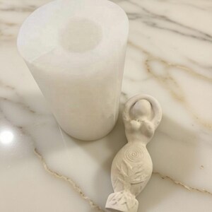 May include: A white, cylindrical candle holder next to a small, off-white statue of a woman with her arms raised. The statue has a spiral design on the torso and a leaf pattern at the base. The items are on a marble surface.