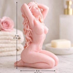 Puede incluir: Una vela de color rosa p&aacute;lido con forma de figura arrodillada con el cabello largo y ondulado y los brazos levantados sobre la cabeza. La figura mide 17,8 cm de alto y 10,2 cm de ancho, seg&uacute;n indican las l&iacute;neas de cota negras en la imagen.