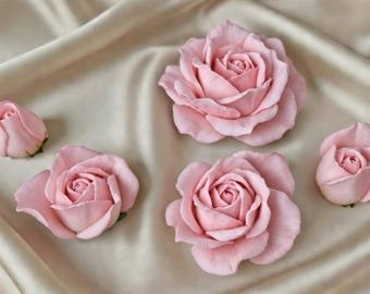 May include: Five light pink rose blooms and buds are arranged on a cream-colored satin fabric. The roses vary in size, with some fully open and others in bud form. The petals have a soft, delicate appearance.