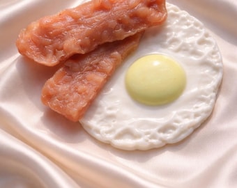 Könnte beinhalten: Ein Stillleben mit einem Spiegelei und zwei Speckstreifen. Das Ei hat eine weiße Basis und ein gelbes Eigelb. Der Speck ist rotbraun. Die Gegenstände sind auf einem Satin-Stoff angeordnet.