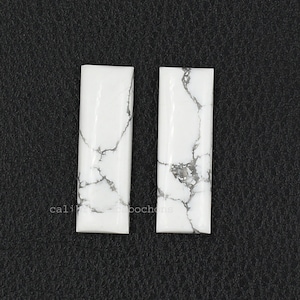 Könnte beinhalten: Zwei weiße Howlith-Cabochons mit grauen Adern. Die Cabochons sind rechteckig und haben eine glatte, polierte Oberfläche.