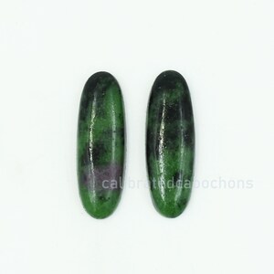 Può includere: Due pietre preziose di forma ovale con un colore di base verde e inclusioni verde scuro e viola. Le pietre preziose sono esposte su sfondo bianco. Il testo "calibratedcabochons" è visibile in basso.