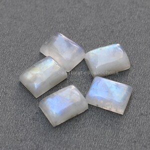 Puede incluir: Cinco cabochones de piedra lunar blanca, cada uno con una forma rectangular y colores iridiscentes azules y blancos. Las piedras están dispuestas en un grupo sobre una superficie gris.