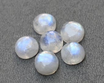May include: Seven round, white moonstone cabochons with iridescent blue and white flashes. The stones are arranged in a loose circle on a gray surface.