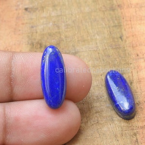 Könnte beinhalten: Zwei ovale Lapislazuli-Cabochons mit goldenen Flocken. Die Cabochons sind tiefblau und haben eine glatte, polierte Oberfläche.