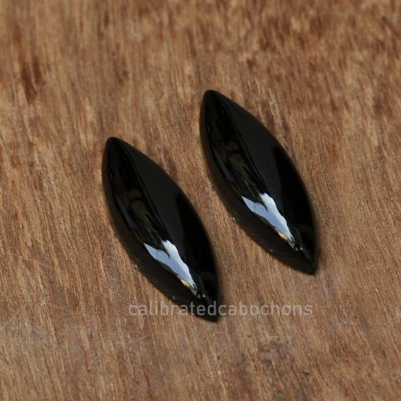 Pu&ograve; includere: Due cabochon neri lucidi a forma di marquise. I cabochon sono lisci e lucenti, con una leggera curvatura. Sono posizionati su uno sfondo di legno marrone.