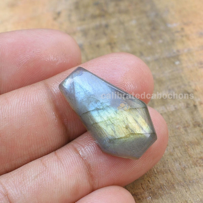 May include: A gray and green labradorite cabochon with iridescent flashes of color. The cabochon is shaped like a teardrop and has a smooth, polished surface.