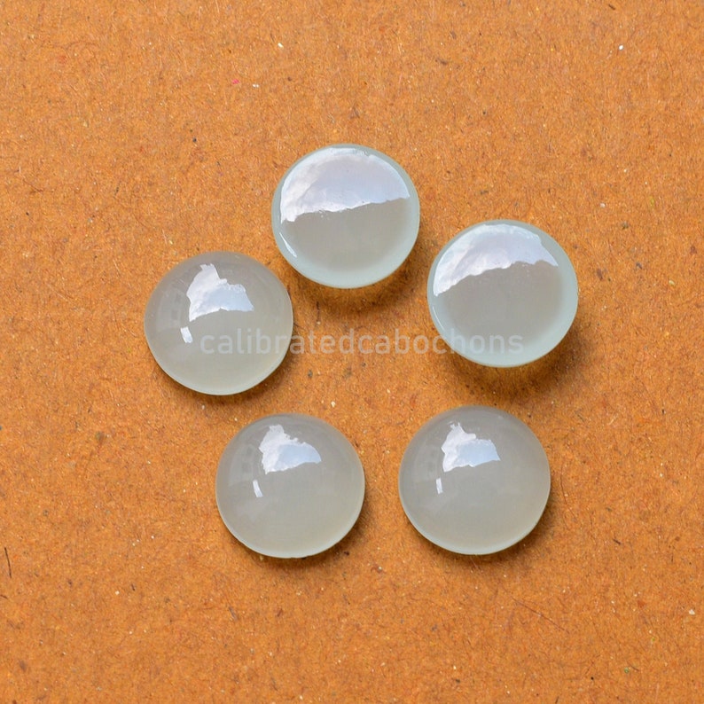 May include: Five white, round, polished cabochons, each with a slightly different shade of white, arranged in a circle on a brown surface.