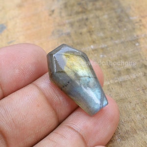 May include: A gray and blue labradorite gemstone cabochon with a pointed, teardrop shape. The stone has a iridescent sheen and is polished to a smooth finish.