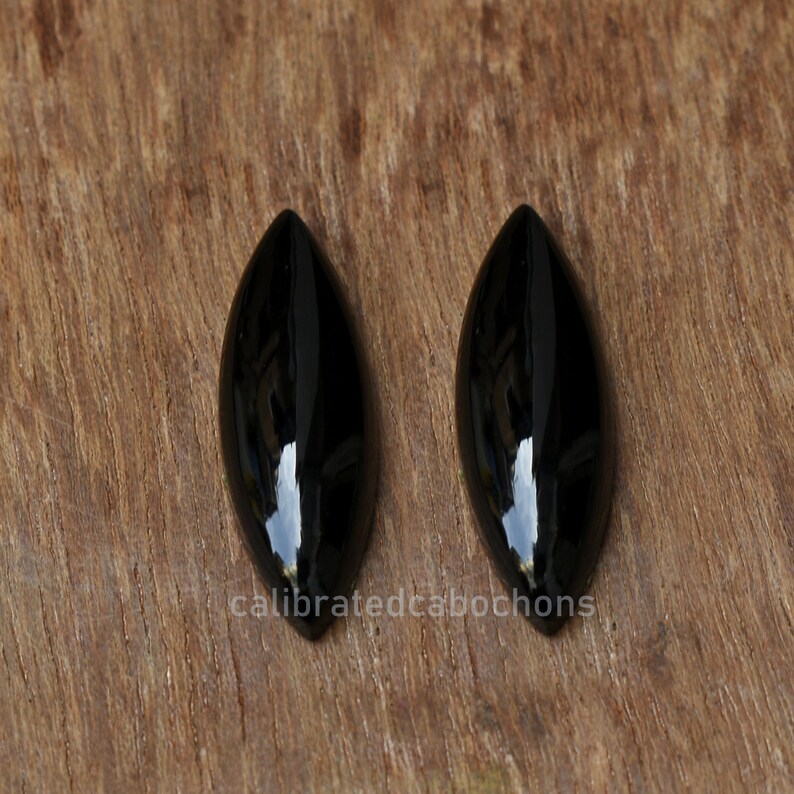 Pu&ograve; includere: Due cabochon neri lucidi a forma di marquise su una superficie di legno.