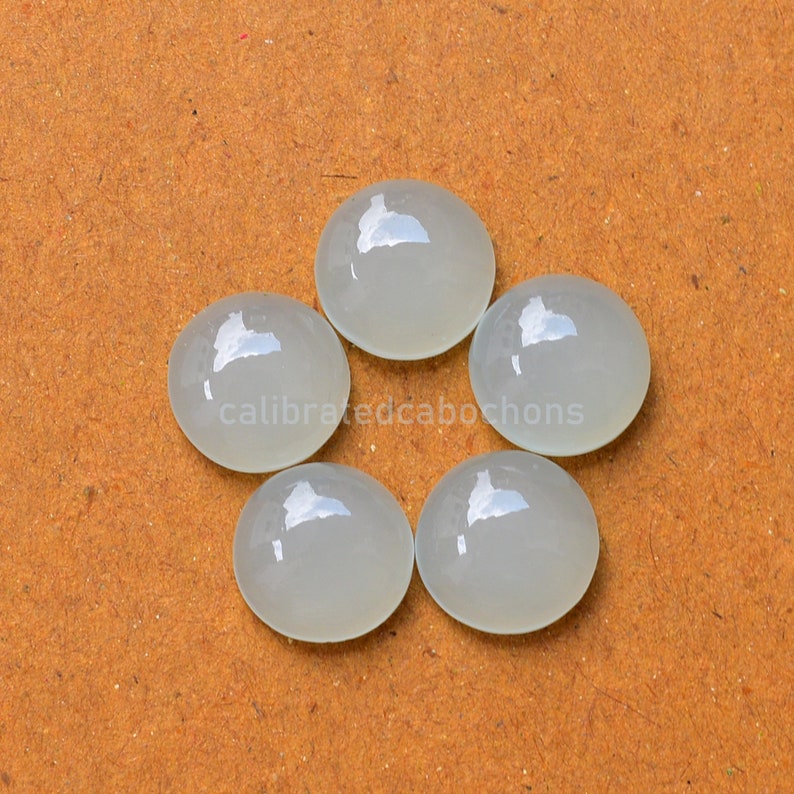 May include: Five round, white, polished cabochons arranged in a circle on a brown surface.