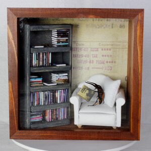 May include: A miniature diorama in a wooden frame features a small bookshelf, a white armchair, and a large insect reading a book. The background includes a vintage document. The bookshelf is filled with tiny books.