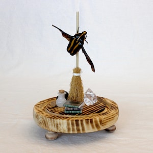 May include: A whimsical wooden display featuring a miniature broom, a butterfly with yellow and black stripes, small books, a crystal, and a tiny bottle with a cork. The display is set on a round, wooden base with three small feet.